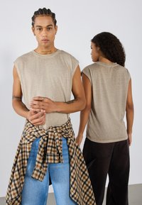 Beige kortermet t-skjorte med tekstur, båret av to modeller. Den ene modellen har en rutete skjorte knyttet i livet over blå jeans.