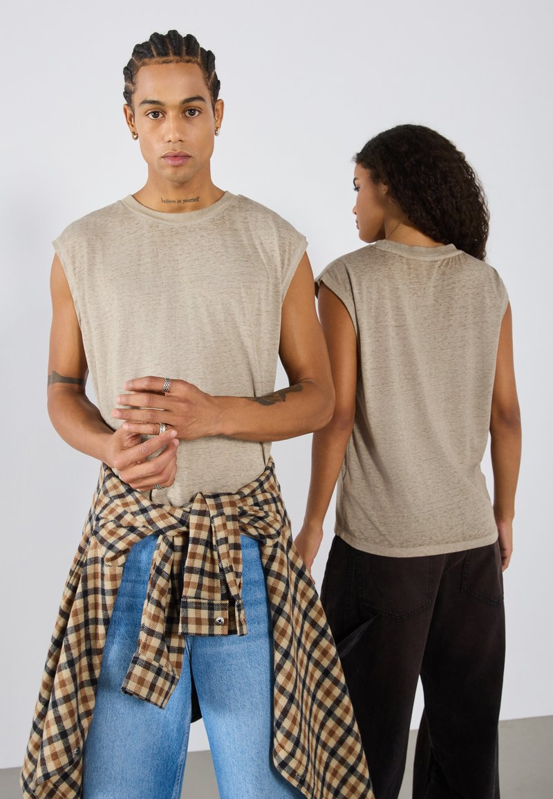 Beige kortermet t-skjorte med tekstur, båret av to modeller. Den ene modellen har en rutete skjorte knyttet i livet over blå jeans.
