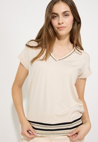 Beige Kurzarm-Oberteil mit V-Ausschnitt und gewelltem Saum sowie einem dekorativen gestreiften Saum in Schwarz, Weiß und Beige. Glatte Textur.