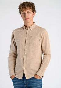 Camisa de botones de manga larga color beige con un tejido texturizado, un bolsillo en el pecho y botones blancos. Combinada con jeans azules.