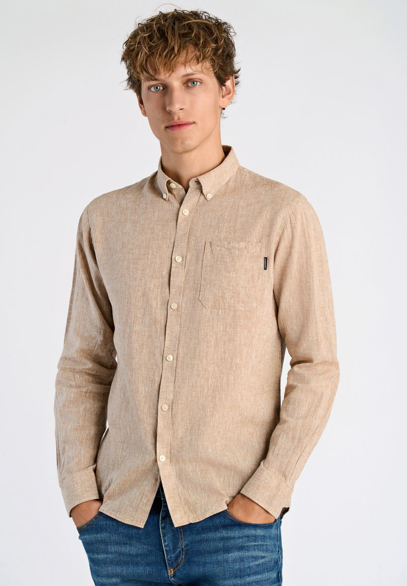 Camicia beige a maniche lunghe con bottoni, dal tessuto texturizzato, con taschino sul petto a sinistra e bottoni bianchi. Abbinata a jeans blu.