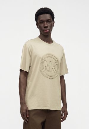 Jeune homme portant un t-shirt beige à manches courtes avec un logo MK embossé, debout devant un fond blanc uni.