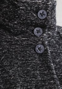 Närbild av ett texturerat svart och vitt tyg med tre mörka knappar och en hög krage, som framhäver plaggets detaljer och design.