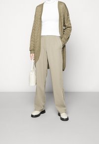 Luftiger olivfarbener Cardigan über einem weißen Rollkragenpullover, beigefarbene weit geschnittene Hose und weiße Handtasche. Die Schuhe haben eine strukturierte schwarze Sohle.