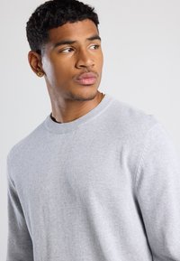 Giovane uomo con i capelli corti e ricci, che indossa un maglione grigio chiaro, un orecchino a vite dorato e una collana, mentre guarda verso sinistra su uno sfondo neutro.