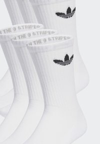 Balti kokvilnas maisījuma zeķes ar ribotu tekstūru, kurās augšā ir melns Adidas logos un pelēks svītrojums ap potīti.