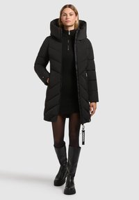 Zwarte pufferjas met een hoge kraag en een oversized capuchon. Beschikt over chevron-quilting, een ritssluiting en een logootje. Gestyleerd met een zwarte gebreide jurk en kniehoge laarzen.