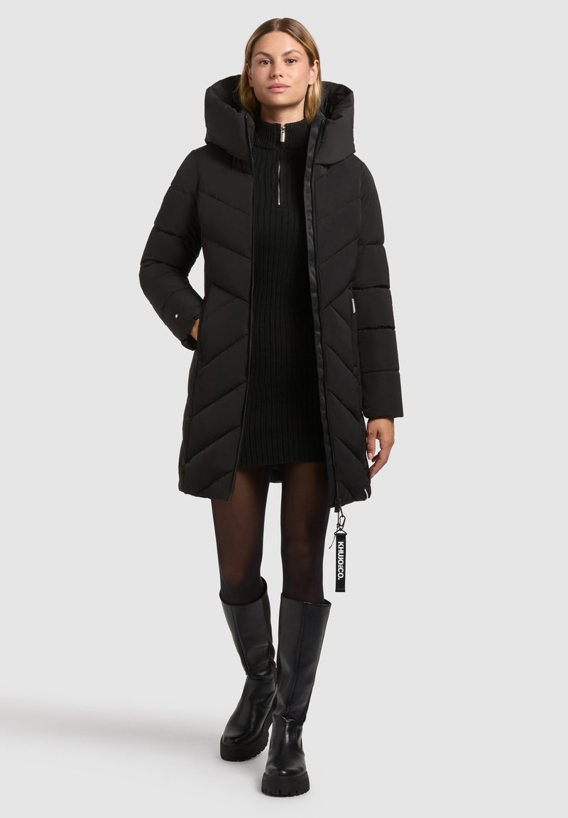 Zwarte pufferjas met een hoge kraag en een oversized capuchon. Beschikt over chevron-quilting, een ritssluiting en een logootje. Gestyleerd met een zwarte gebreide jurk en kniehoge laarzen.