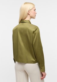 Blusa in satin verde oliva con colletto, dettagli delle cuciture visibili e polsini con bottoni. Il tessuto presenta una texture liscia e una finitura leggermente lucida.