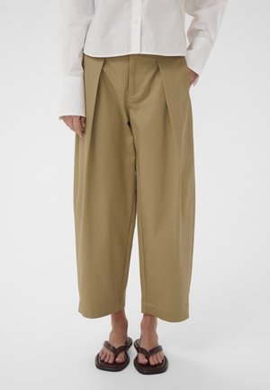 Személy bézs, széles szárú, rövidnadrágos nadrágot, fehér hosszú ujjú inget és sötétbarna, négyzet alakú orrú szandált visel, fehér háttér előtt áll.