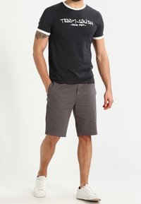 Homme portant un T-shirt noir avec motif, short gris longueur genou, baskets blanches, debout avec une main dans la poche devant un fond neutre.