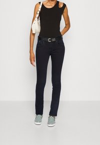 Černý bezrukávový top, tmavě modré džíny s páskem a zelené kostičkované mokasíny. Bílé kabelka s popruhem. Jednoduché pozadí.