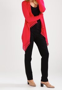 Světle růžový kardigan s krajkovým lemem na spodním okraji, v kombinaci s černými přiléhavými kalhotami a béžovými botami na blokovém podpatku. Hladká textura, otevřený přední design.