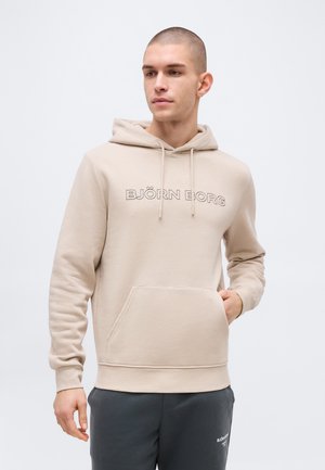 Άνδρας που φοράει μπεζ φούτερ Björn Borg με τσέπη εμπρός και κορδόνια, σε συνδυασμό με σκούρο γκρι φόρμα Björn Borg, στέκεται μπροστά σε λευκό φόντο.