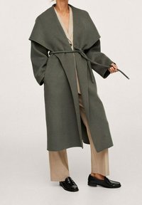 Lang, olijfgroene jas met brede revers en een strikceintuur, gemaakt van textuurmateriaal, gedragen over een beige outfit en zwarte instapschoenen.