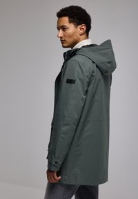 Green waterproof jacket with a hood, front zipper, large pockets, and black accents. Smooth texture, mid-length design, and casual fit.