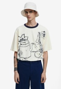 T-shirt crema a maniche corte con disegni in linea nera di un annaffiatoio, fiori e frutta; abbinato a pantaloni blu navy.