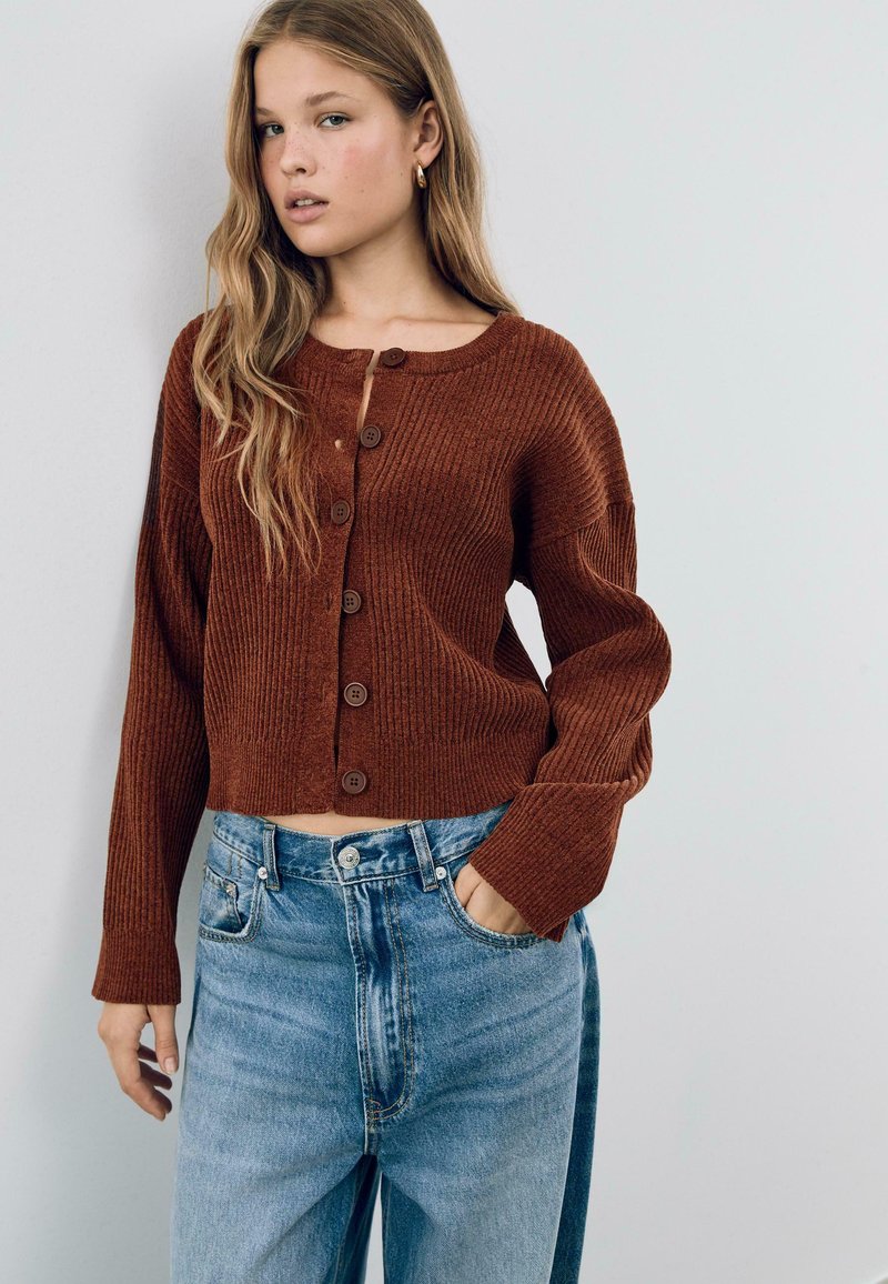 Cardigan court côtelé marron avec sept boutons, col rond, et manches longues, associé à un jean en denim bleu clair taille haute.