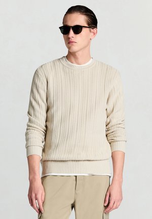 Pull beige en tricot avec poignets et ourlet à côtes, présentant un motif texturé. Porté sur une chemise blanche, accompagné de lunettes de soleil noires.