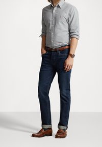 Niebieskie dżinsy o slim fit, zestawione z jasnoszaryą koszulą zapinaną na guziki. Brązowy skórzany pasek i brązowe buty eleganckie. Podwinięte rękawy koszuli.