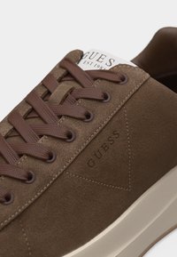 Brūnas zamšādas sporta kurpes ar brūnām šņorēm, sānos iespiests "GUESS" logotips, baltu ārpusi un daļēju mēles etiķeti ar uzrakstu "GUESS EST 1981".