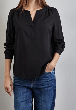 Vrouw die een zwart blouse met lange mouwen en een V-hals draagt, met een blauwe spijkerbroek, staand met één hand in haar zak tegen een effen muur.