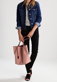 Giacca di jeans, top grigio a righe, pantaloni neri, che tiene una borsa tote rosa chiaro con due manici e tacchi aperti neri.