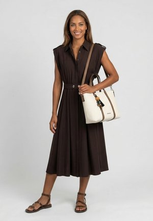 Femme souriante, portant une robe midi marron foncé sans manches et des sandales marron, tenant un sac à main blanc avec des anses marron et des perles décoratives.