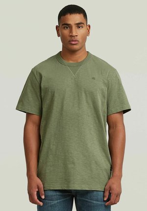 Uomo con capelli corti che indossa una t-shirt verde oliva a maniche corte semplice e jeans blu, in piedi davanti a uno sfondo neutro.