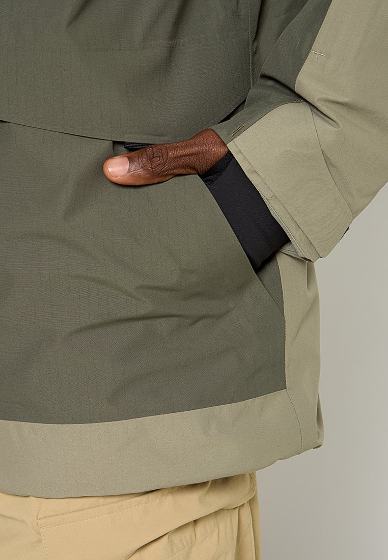 Chaqueta verde oliva con acentos en un verde claro contrastante, que presenta un bolsillo lateral y una tela texturizada. La manga tiene un acabado liso.