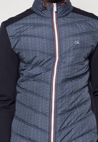 Puffer jacket with herringbone pattern in navy and gray fabric. Features quilted body, navy sleeves, and orange-accented zipper.