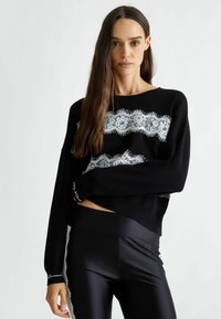 Maglione a maglia nera con un motivo in pizzo bianco sul petto, dotato di maniche lunghe e orlo corto. Abbinato a leggings neri lucidi.