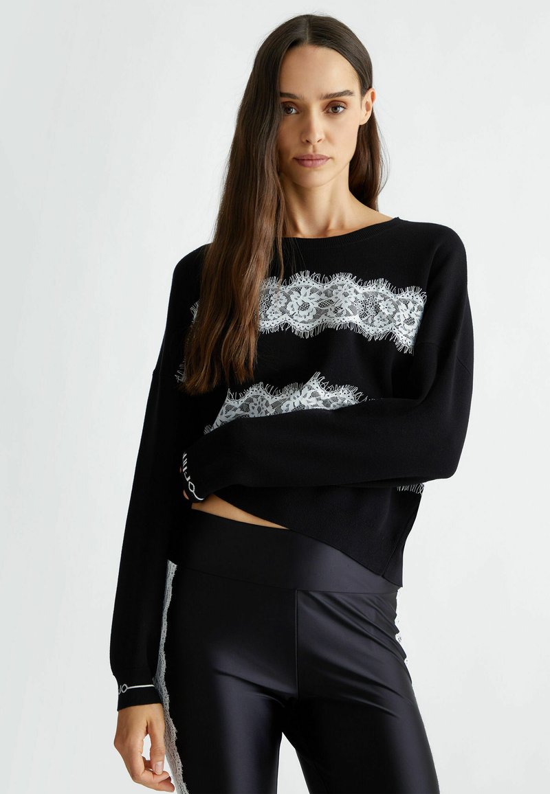 Maglione a maglia nera con un motivo in pizzo bianco sul petto, dotato di maniche lunghe e orlo corto. Abbinato a leggings neri lucidi.