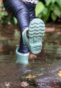 Bottes de pluie en caoutchouc vert menthe avec des semelles texturées marchant à travers une eau peu profonde, entourées de pierres humides et de feuilles tombées.