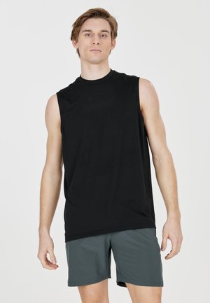 Ung mand iført en ærmeløs sort skjorte og grå shorts står mod en ensfarvet lys baggrund.