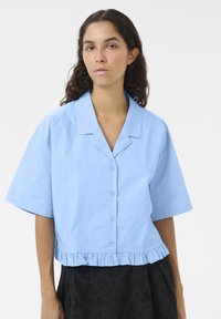 Camicia cropped di colore azzurro chiaro con chiusura frontale a bottoni, maniche corte e orlo arricciato, realizzata in tessuto morbido di colore uniforme.