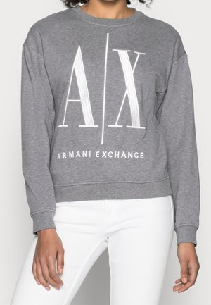 Osoba nosząca szarą bluzę z długim rękawem z dużym białym tekstem "A|X" i "Armani Exchange", w połączeniu z białymi spodniami.