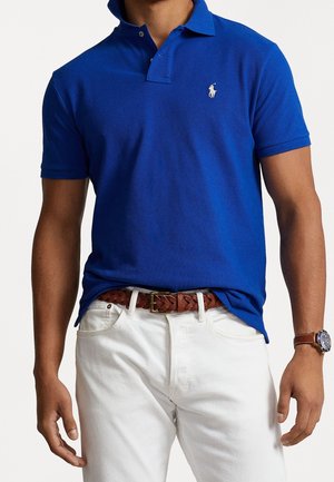 Blaues Poloshirt mit Kragen, auf dem die Brust ein Logo aufgedruckt ist. Getragen mit weißen Jeans und einem braunen geflochtenen Gürtel. Silberne Uhr am Handgelenk.