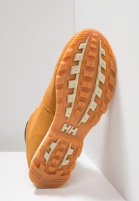 Bruine wandelschoen zool met diep profiel, oranje onderkant, witte accenten en een logo. Zichtbare stiksels accentueren de ontwerpelementen.