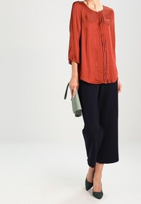 Rostfarvet silke bluse med folder, parret med marineblå culottes og en grøn clutch. Har trekvartærmer og en rund halsudskæring.