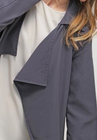 Szara lekka kurtka z asymetrycznym kołnierzem, długimi rękawami i miękką fakturą, noszona na jasnej bluzce.