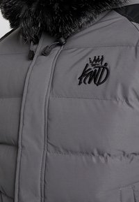 Grå puffjacka med en glänsande svart pälskrage, framdragkedja och en svart broderad logotyp på bröstet. Slät, vadderad textur.