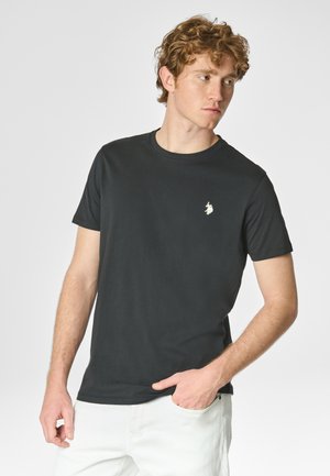 T-shirt nera in cotone con maniche corte, scollo rotondo e un piccolo logo ricamato sul petto. Indossata con pantaloni bianchi.