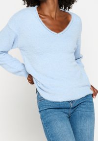 Pull col en V bleu clair en matière douce et duveteuse, à manches longues et coupe décontractée. Surface texturée avec un léger éclat.