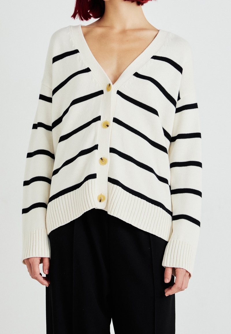 Femme portant un cardigan blanc avec des rayures horizontales noires et quatre boutons, associé à un pantalon noir ample.