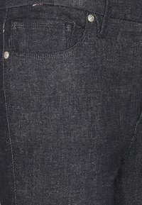 Vaqueros de mezclilla oscura con una superficie texturizada, que incluyen un pequeño bolsillo delantero, herrajes plateados y un detalle de costura en contraste.