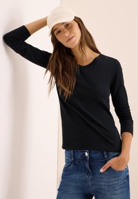 Svart långärmad tröja med rund halsringning, matchad med blå denimjeans. En ljust färgad keps bärs för att ge en avslappnad accent.