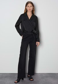 Schwarze Satin-Wickelbluse mit langen Ärmeln, kombiniert mit hochtaillierten schwarzen weiten Jeans, die über ein dezentes Seitennahtdetail verfügen. Schwarze Sandalen mit offenen Zehen.