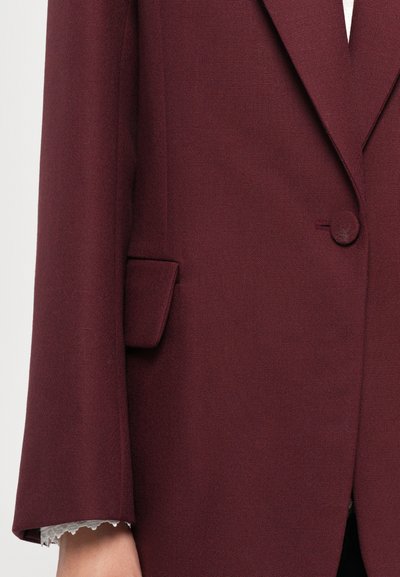 Woman’s hand visible wearing burgundy blazer with flap pocket and fabric-covered button, white lace shirt cuff peeking out.