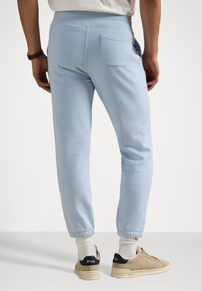 Pantalons de jogging bleu clair avec taille élastique et poignets élastiques. Comprend une poche arrière et une texture lisse. Portés avec des baskets et des chaussettes neutres.